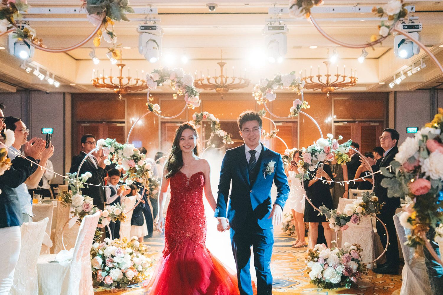 Bride in red gown and groom in blue suit walk hand-in-hand through a grand wedding reception hall with floral decor.