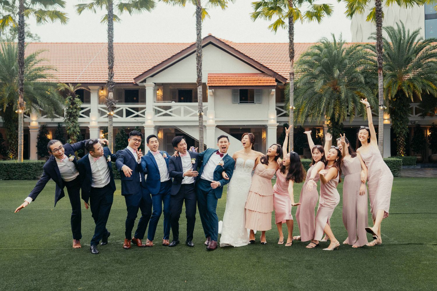 Wedding party celebrating outdoors on lawn with palm trees, groomsmen in navy suits and bridesmaids in blush pink dresses.