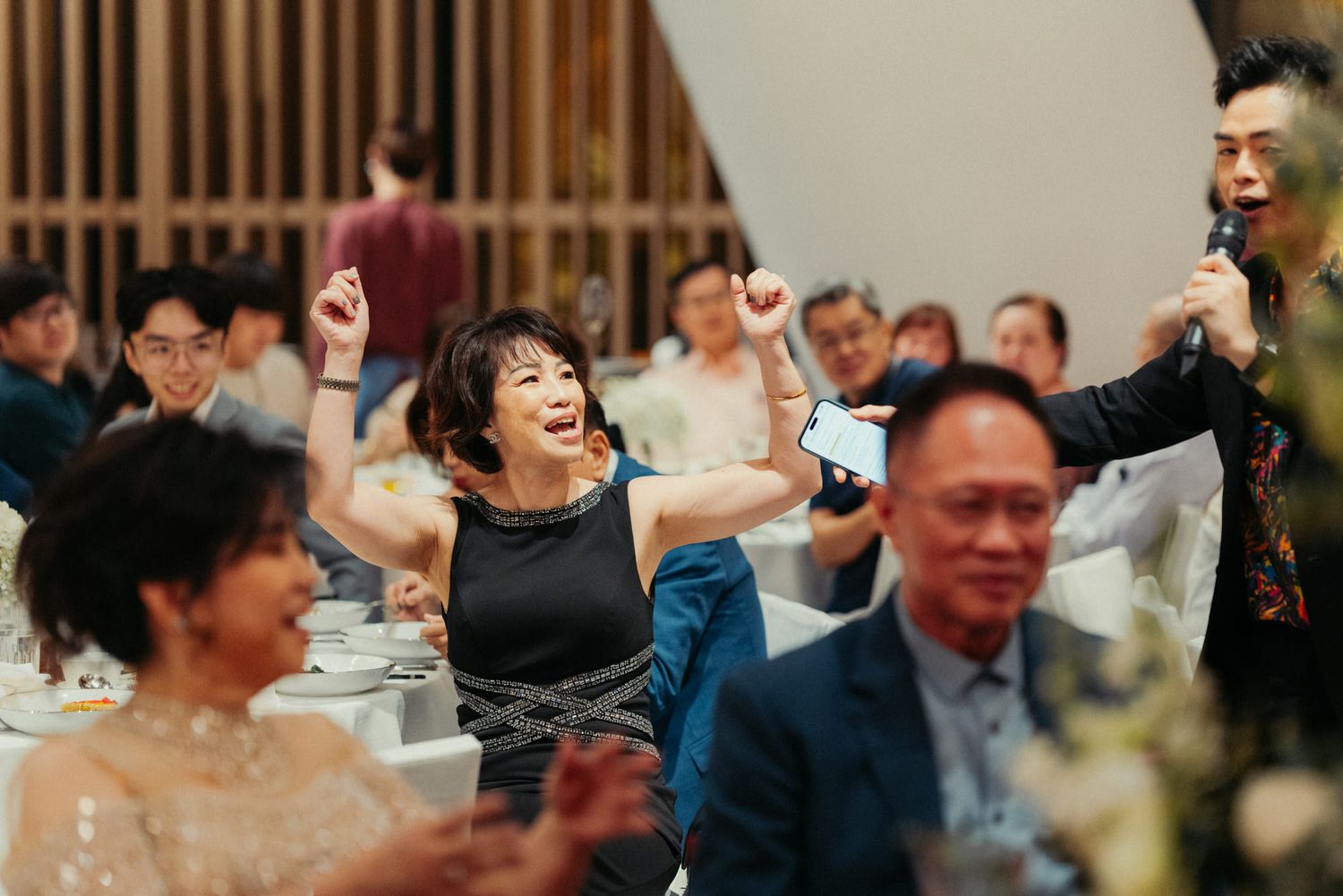 Joyful wedding guest raises arms in celebration during reception dinner with guests and speaker visible.