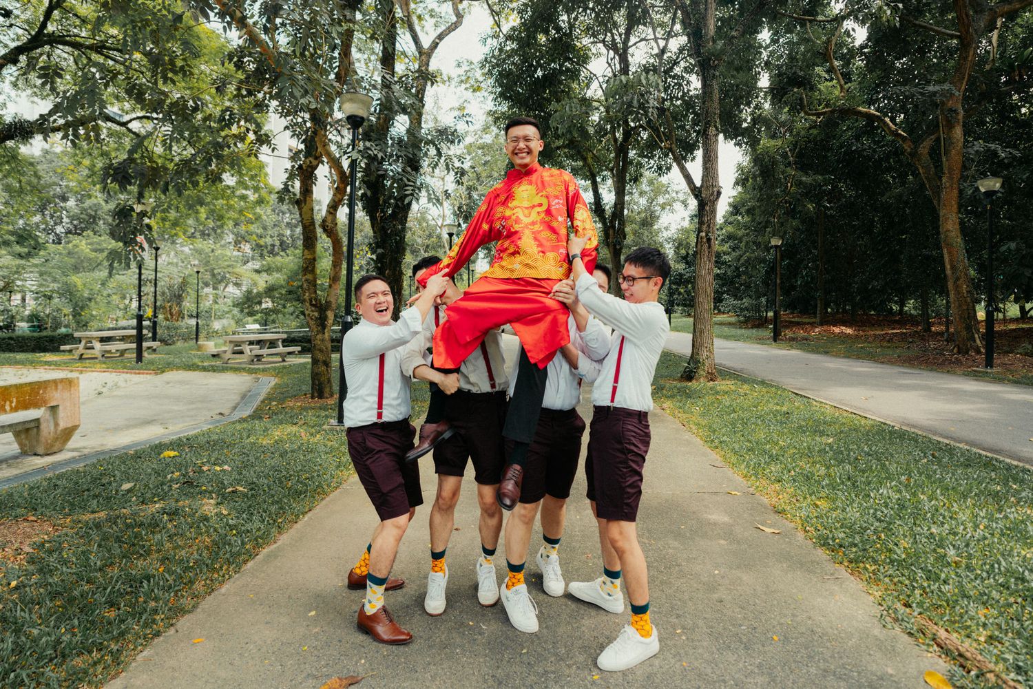 Group of young men in matching uniforms lifting a smiling man in red traditional attire in a park.