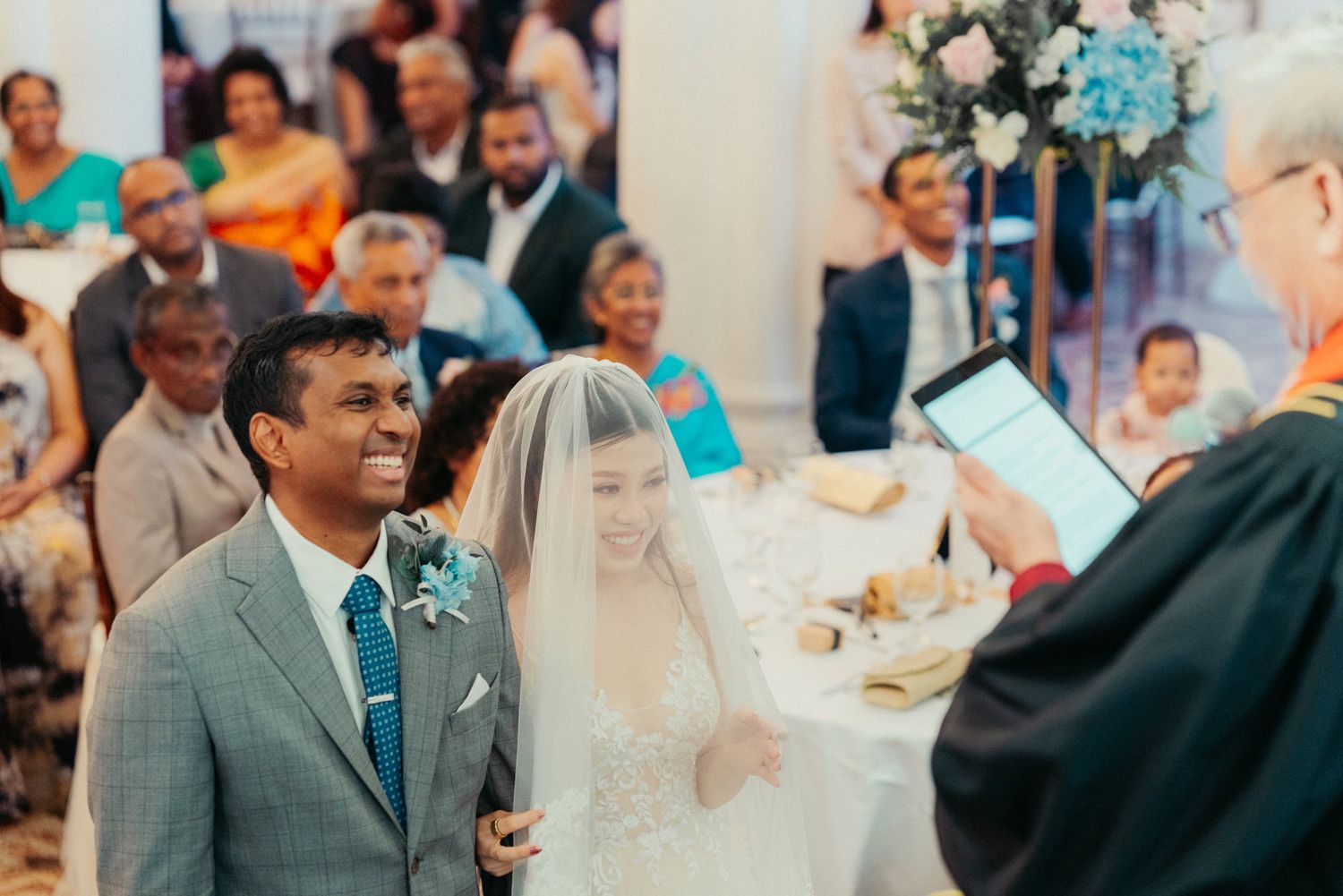 Interracial couple exchange vows before officiant at elegant church wedding ceremony with guests seated behind.