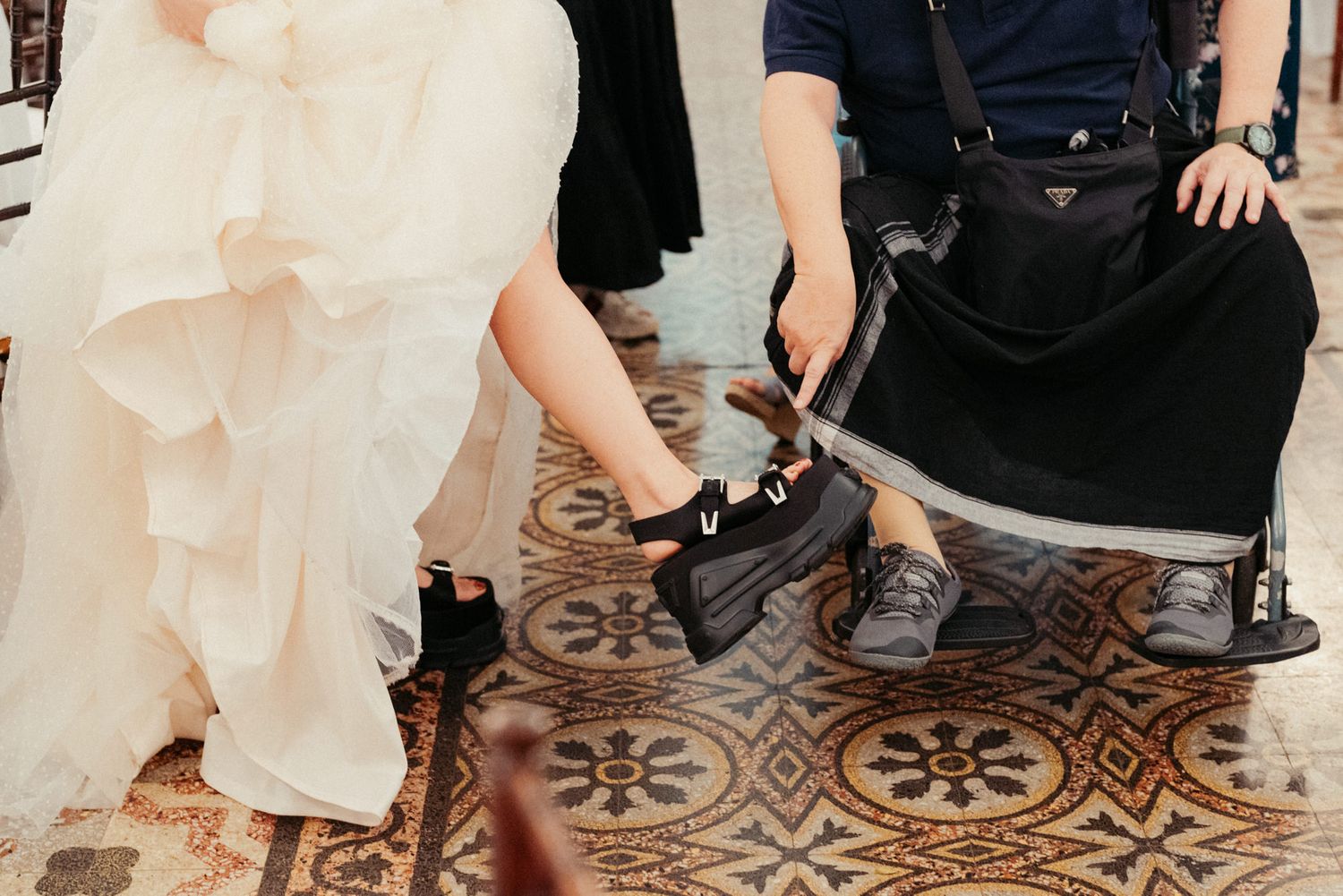 Bride in white gown wearing black platform shoes beside a Prada bag on a patterned tile floor.