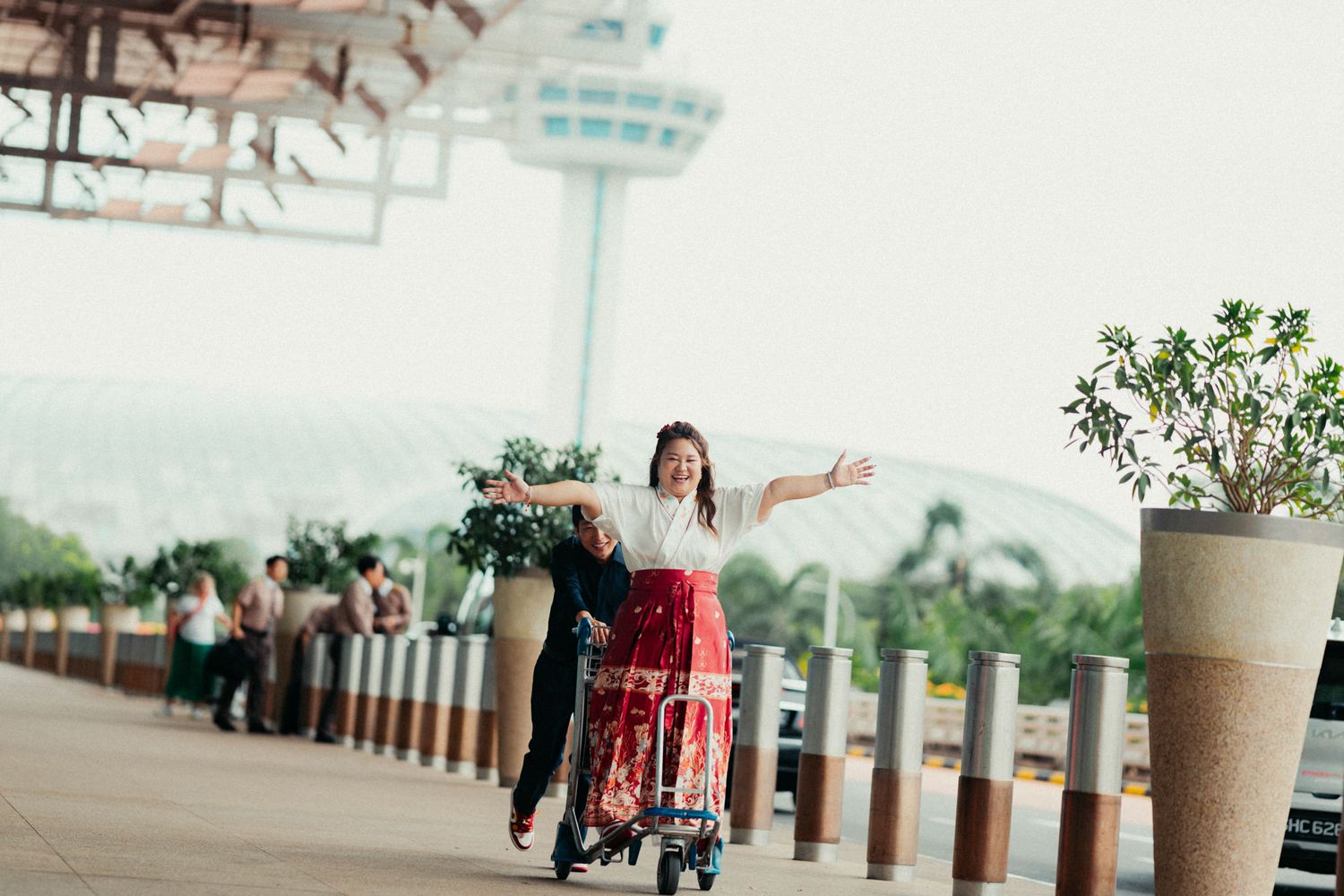 A joyful couple at an airport, woman in red skirt riding a luggage cart pushed by her partner outdoors.