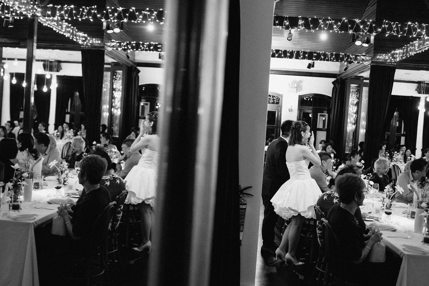 A black and white photo shows a formal dining event with people seated at tables under elegant lighting.