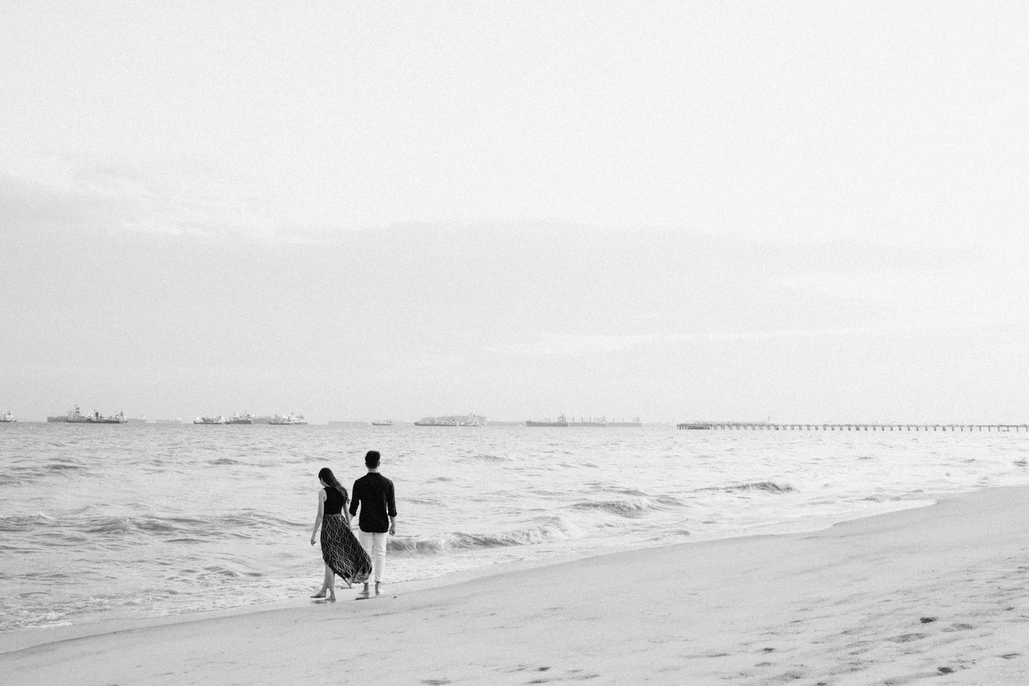 Distant view of couple walking along empty beach with ships on horizon.