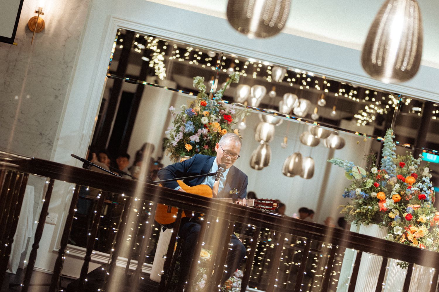 Man in suit reading at elegant restaurant with fairy lights, floral arrangements, and pendant lamps.