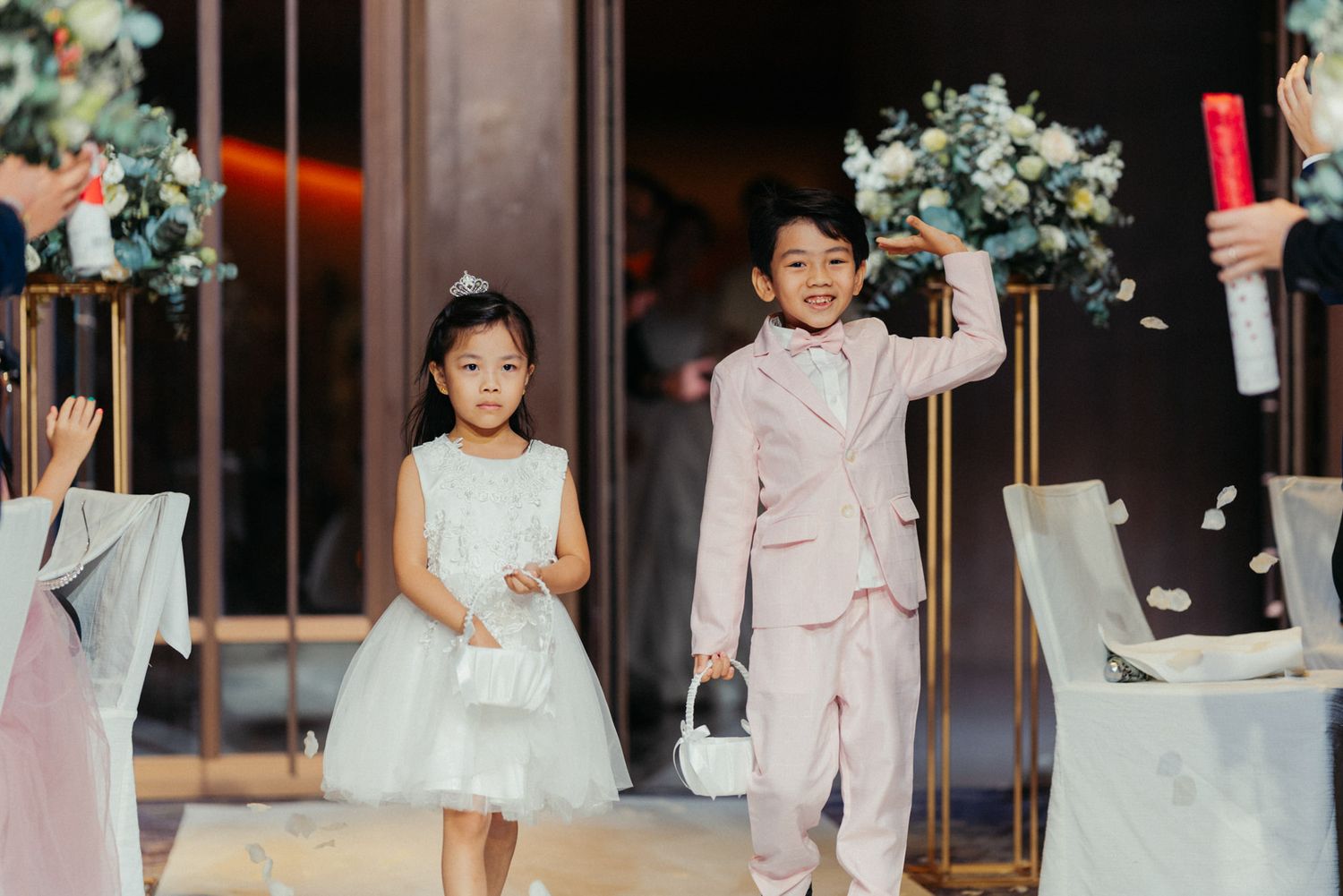 Two children in formal attire walk down a wedding aisle, a girl in white dress and boy in pink suit carrying flower basket.