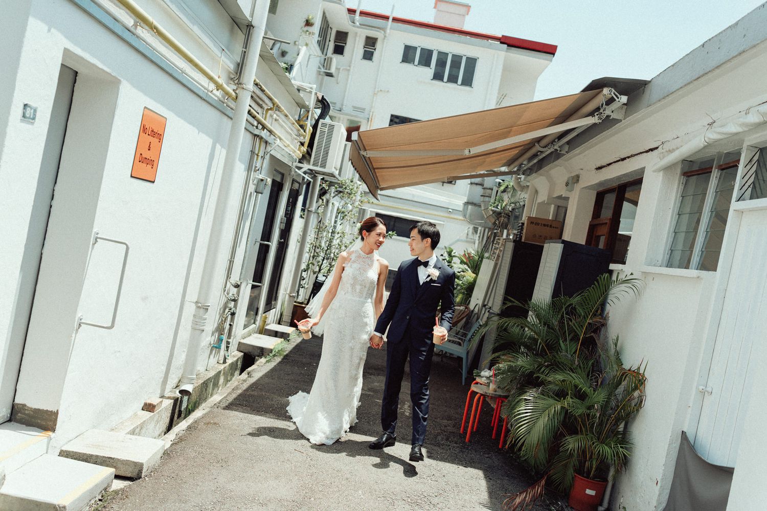 A couple in wedding attire walks hand in hand along a charming outdoor alley with white buildings and lush plants.