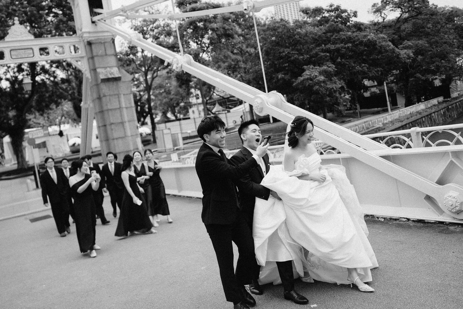 Black and white photo of wedding party walking and posing playfully on a decorative bridge.