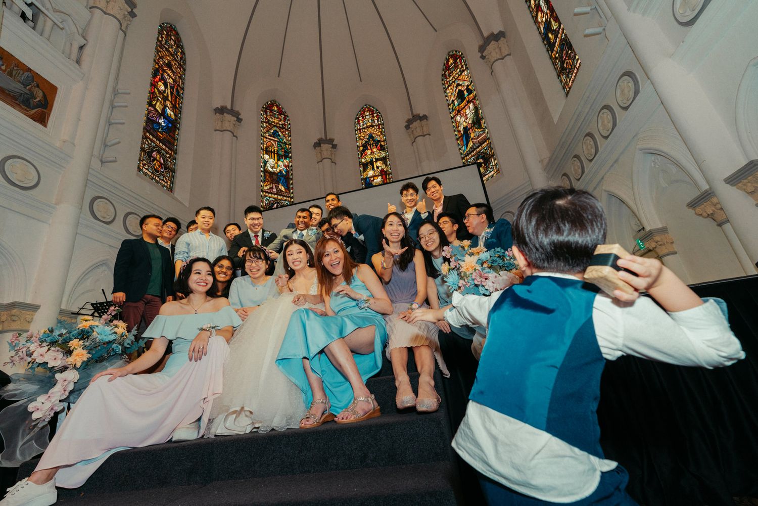 Wedding party poses for group photo inside elegant church with stained glass windows.