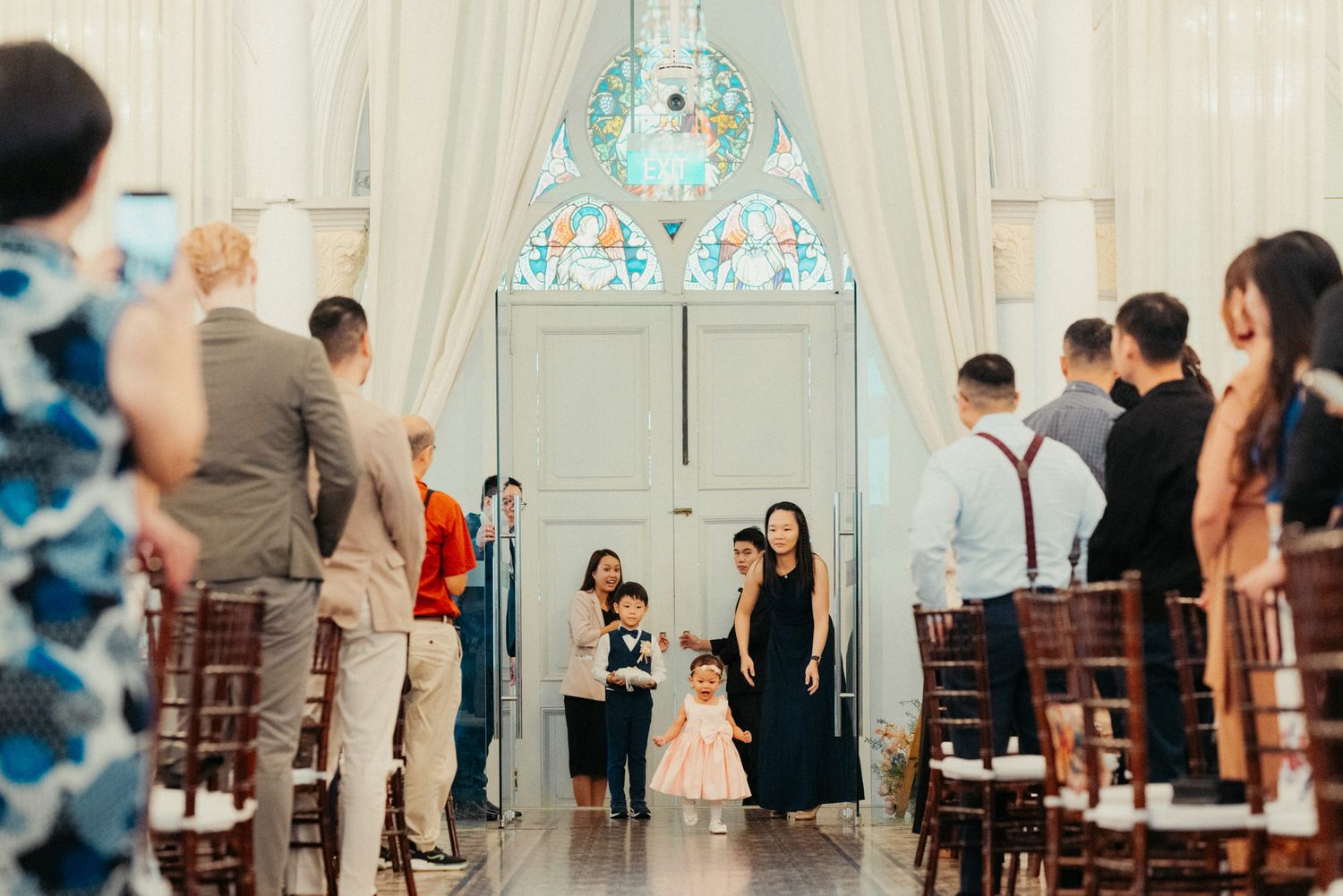 A wedding ceremony aisle in an elegant white chapel with stained glass windows, guests standing as a woman and children walk forward.