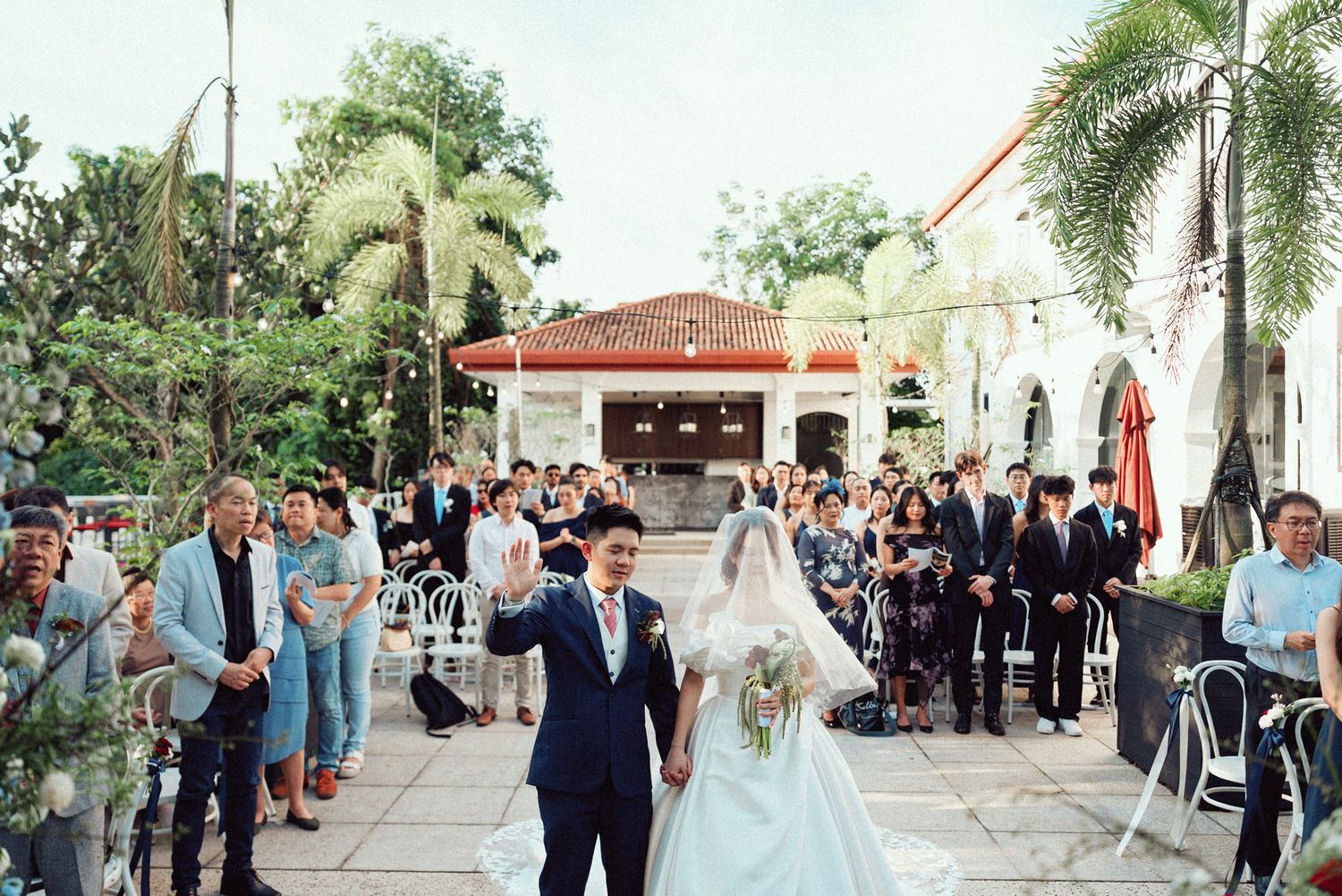 A wedding couple walks down an outdoor aisle lined with palm trees and guests at a Spanish-style venue.
