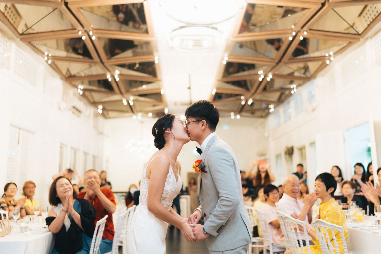 Bride and groom share first kiss at elegant wedding reception as guests applaud in bright white venue.