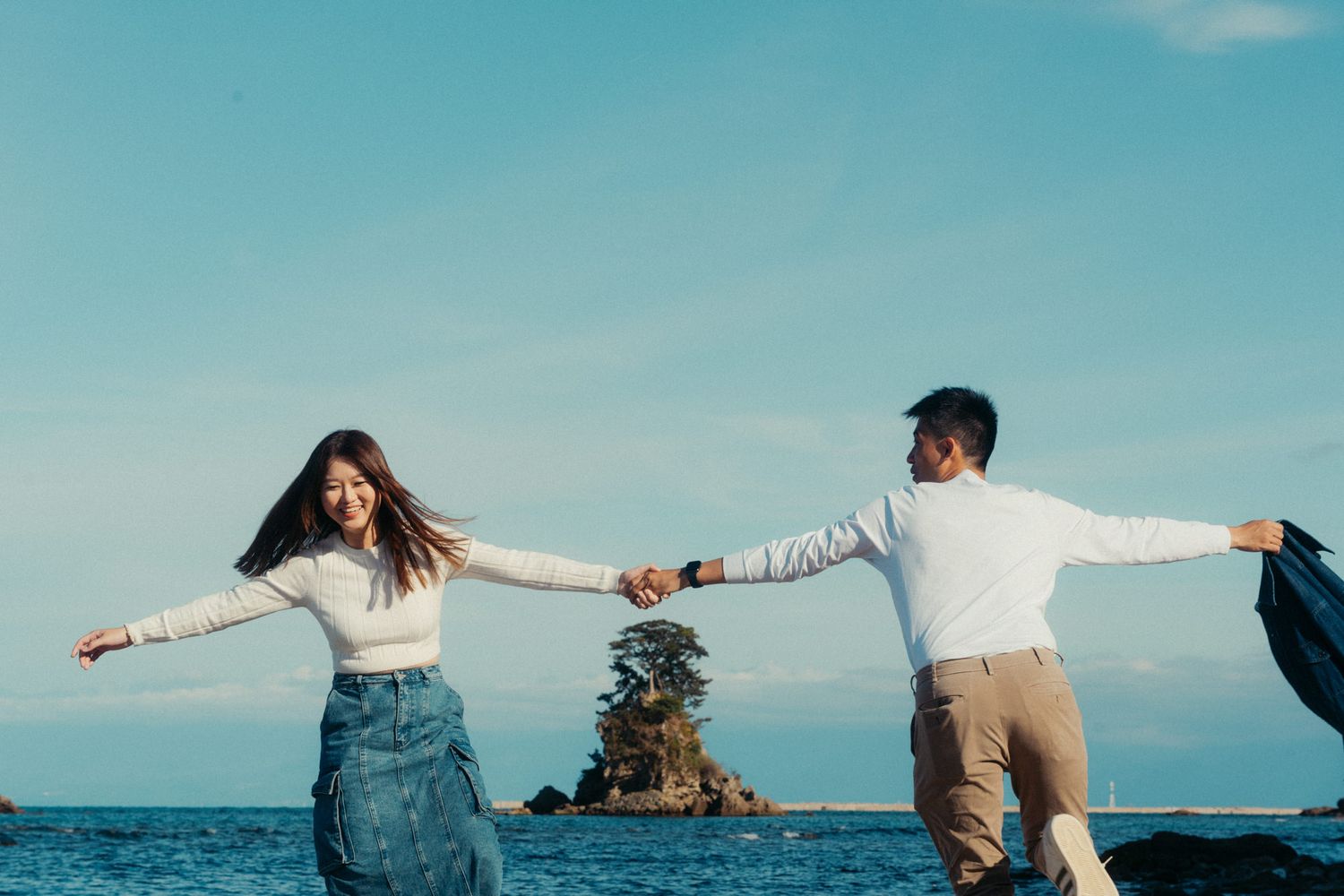 A couple holding hands with arms outstretched against an ocean backdrop with a small island.
