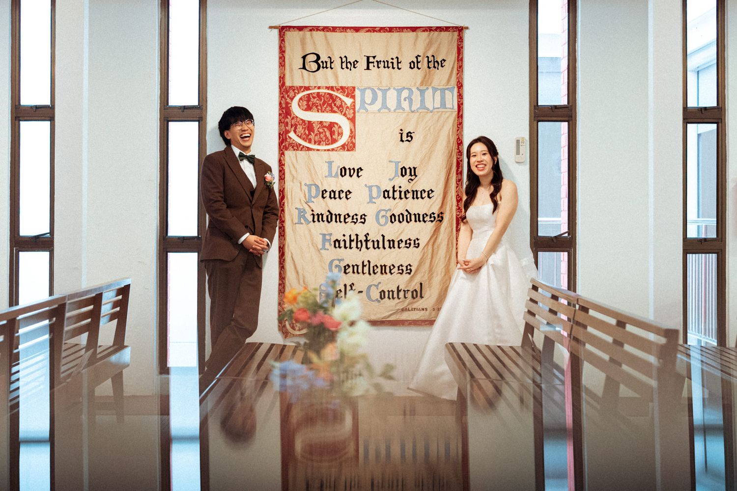 Couple poses in wedding attire before a Fruit of the Spirit banner in a bright, modern chapel.