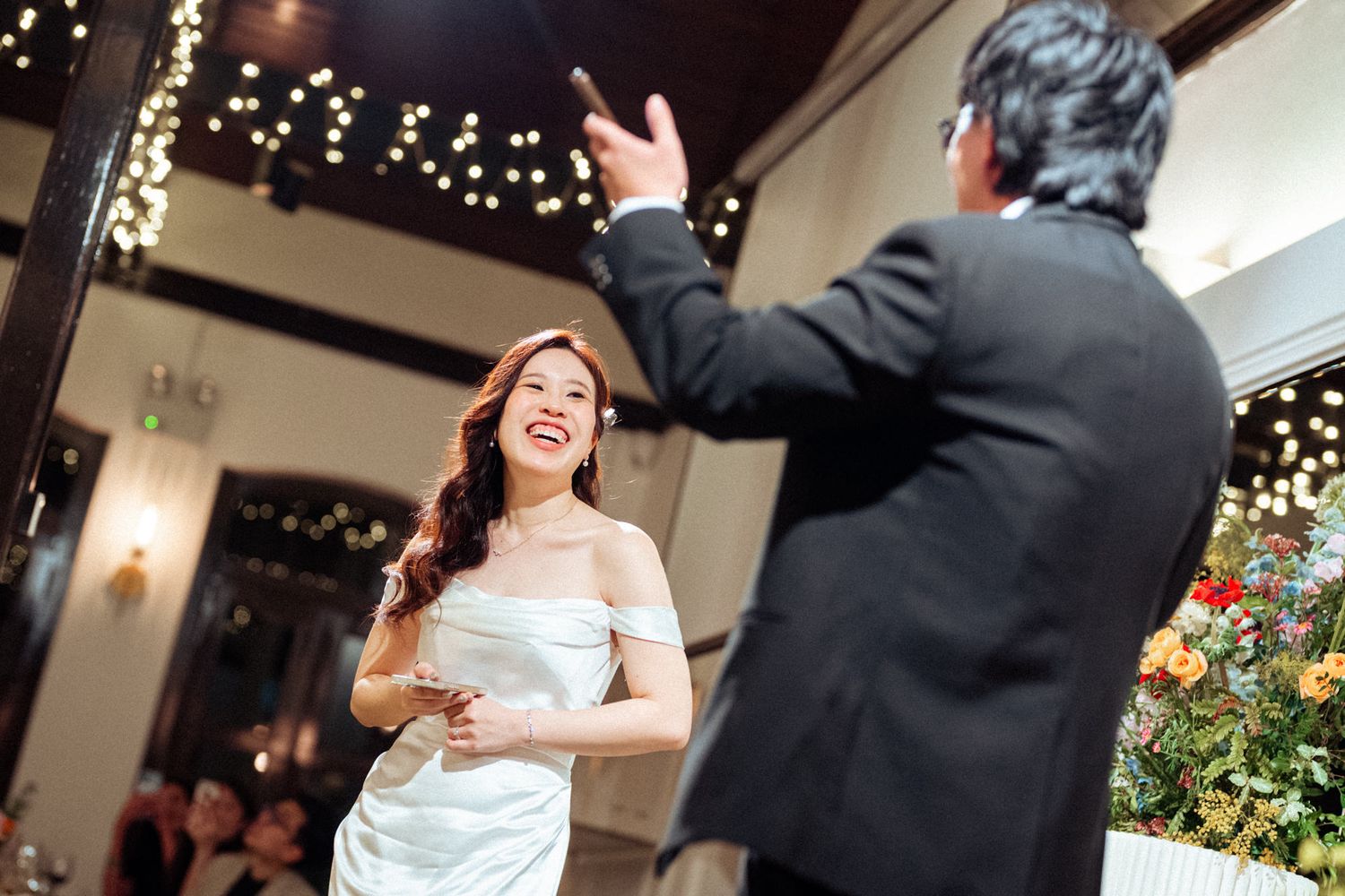 Bride in white off-shoulder gown laughing joyfully during first dance at elegant wedding reception with string lights.