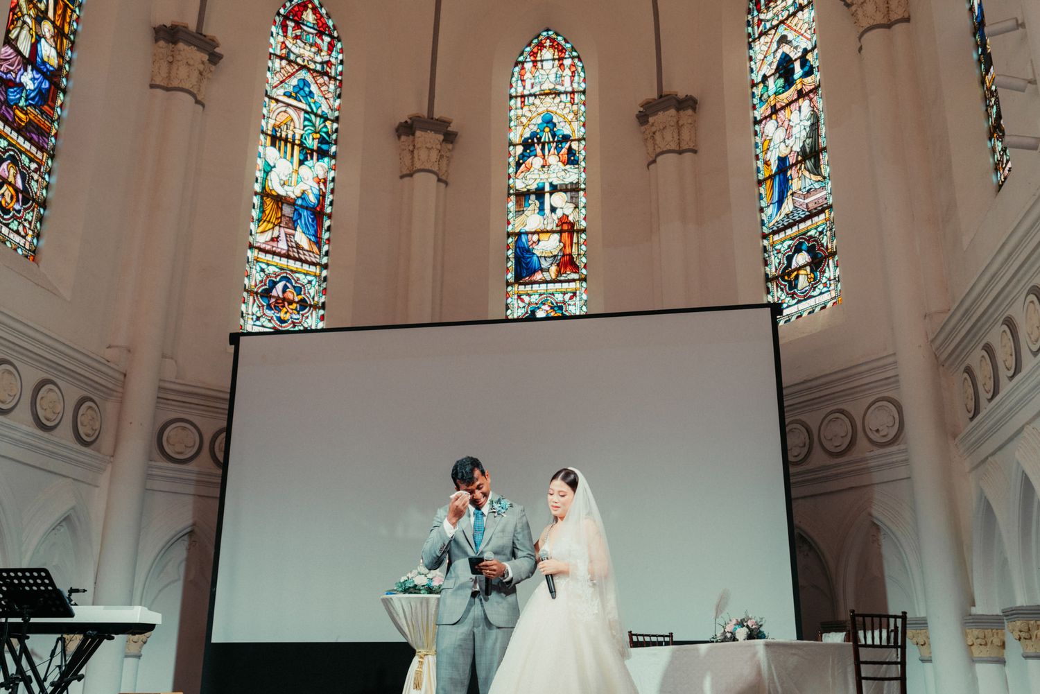 Bride and groom stand together at church altar with colorful stained glass windows behind them.