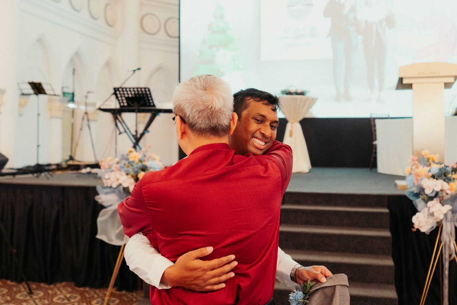 Two men sharing a warm embrace at a formal event, wearing red jackets in an elegant venue.