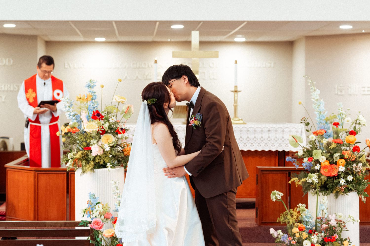 Couple shares first kiss at church wedding altar with colorful floral arrangements and officiant reading ceremony.