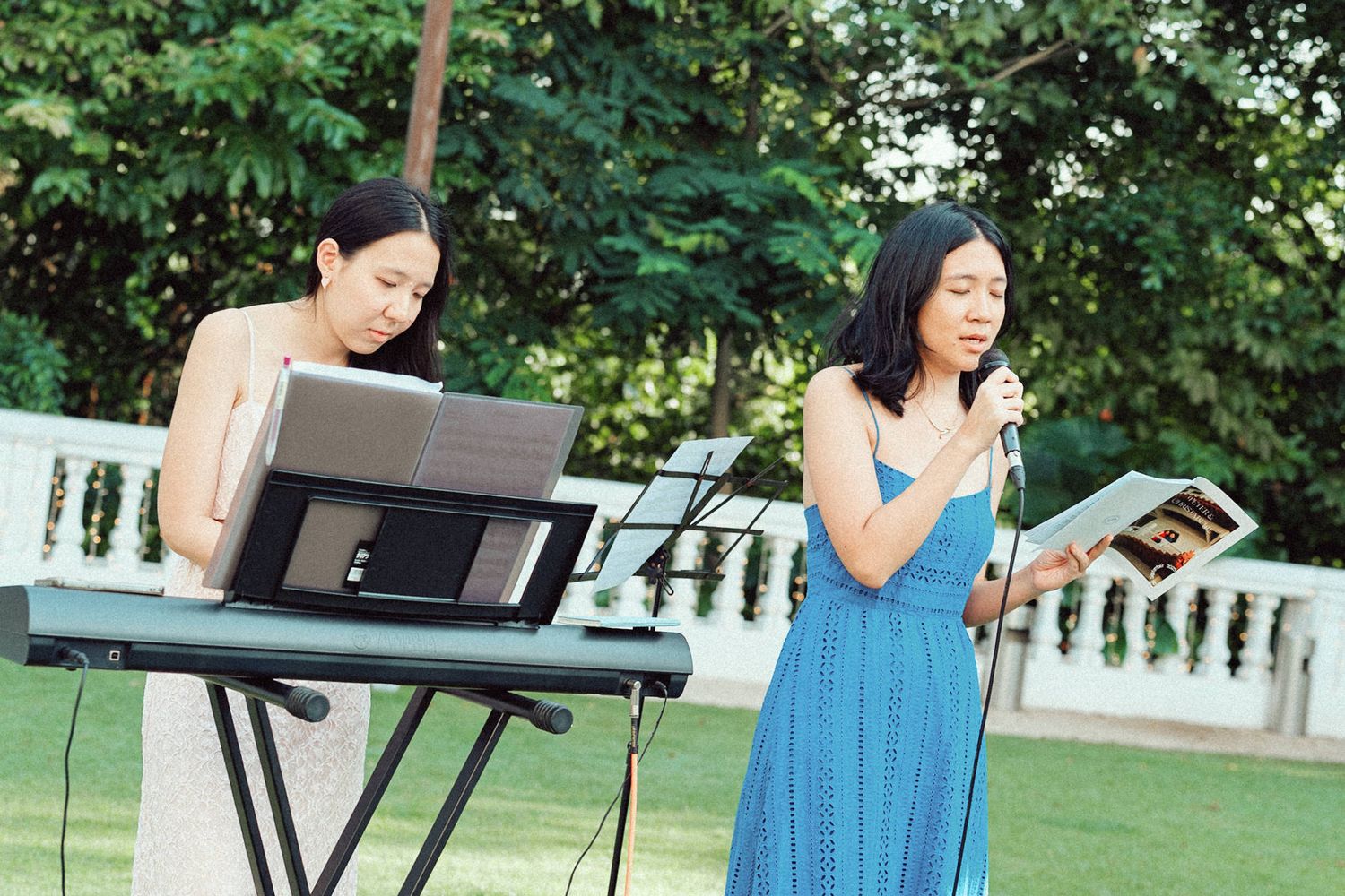 Two performers entertain outdoors, with one playing keyboard and another singing in a blue dress by a white railing.