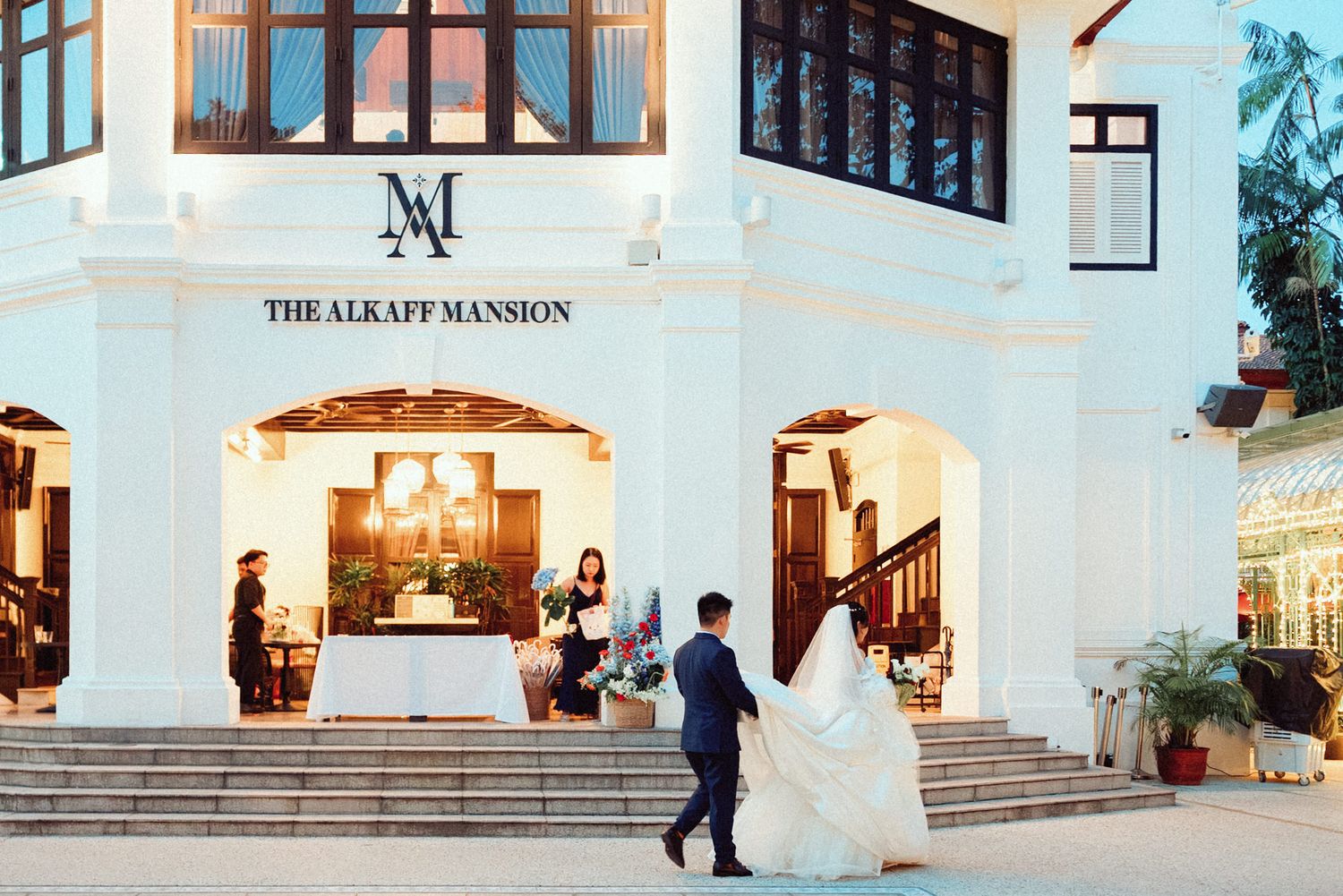 Elegant white mansion wedding venue exterior with illuminated entrance and grand staircase at dusk.