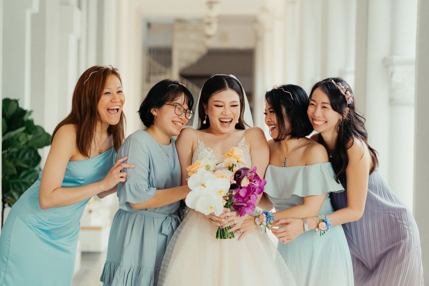 Bride in white gown laughing with four bridesmaids in blue dresses, holding colorful bouquet at elegant wedding venue.