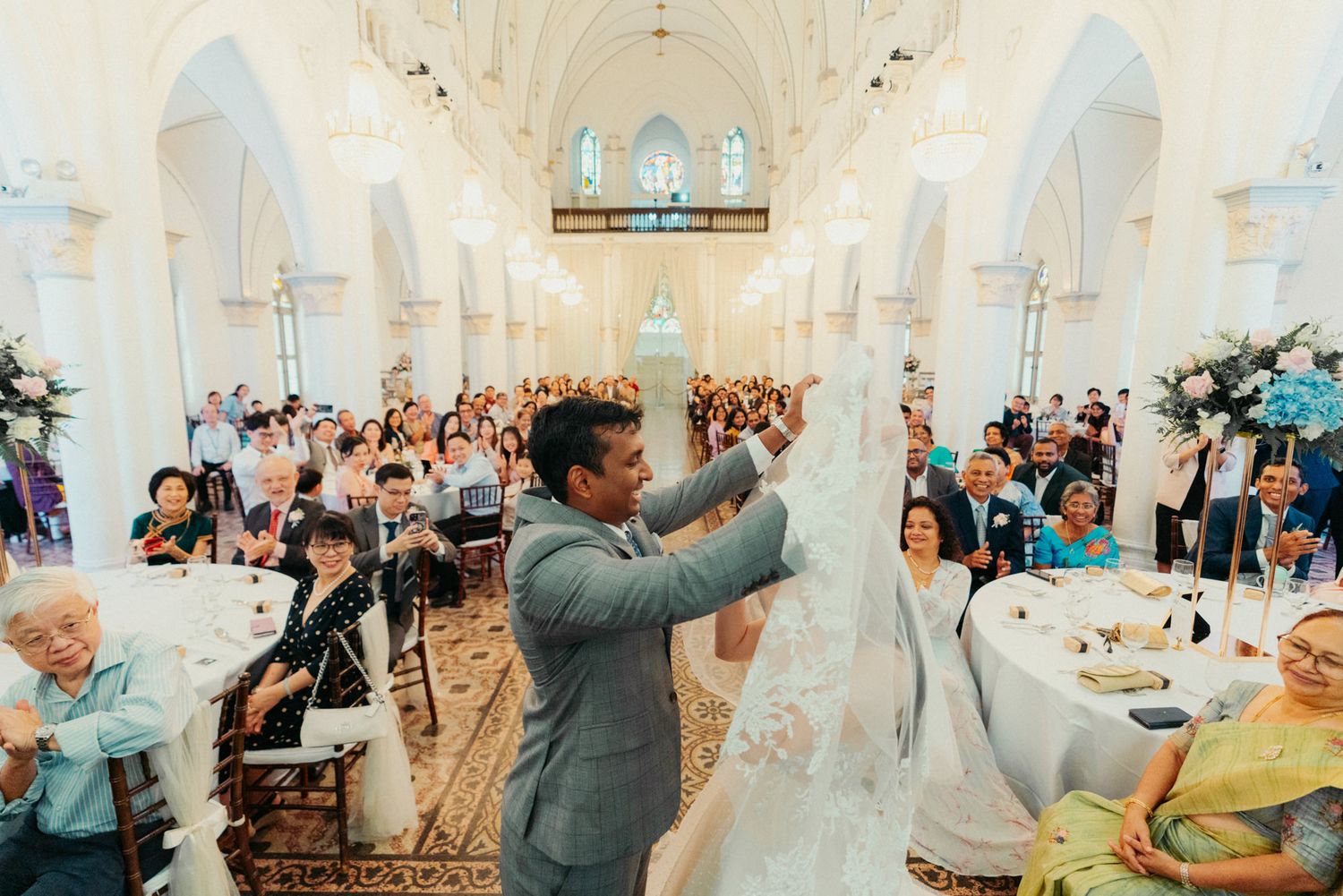Groom lifts bride's veil in elegant church wedding ceremony with guests seated at decorated reception tables.