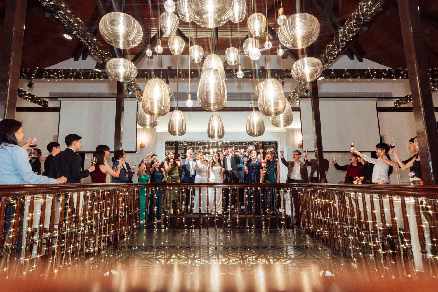 Wedding party celebrates on a grand staircase under ornate hanging lanterns in an elegant venue.