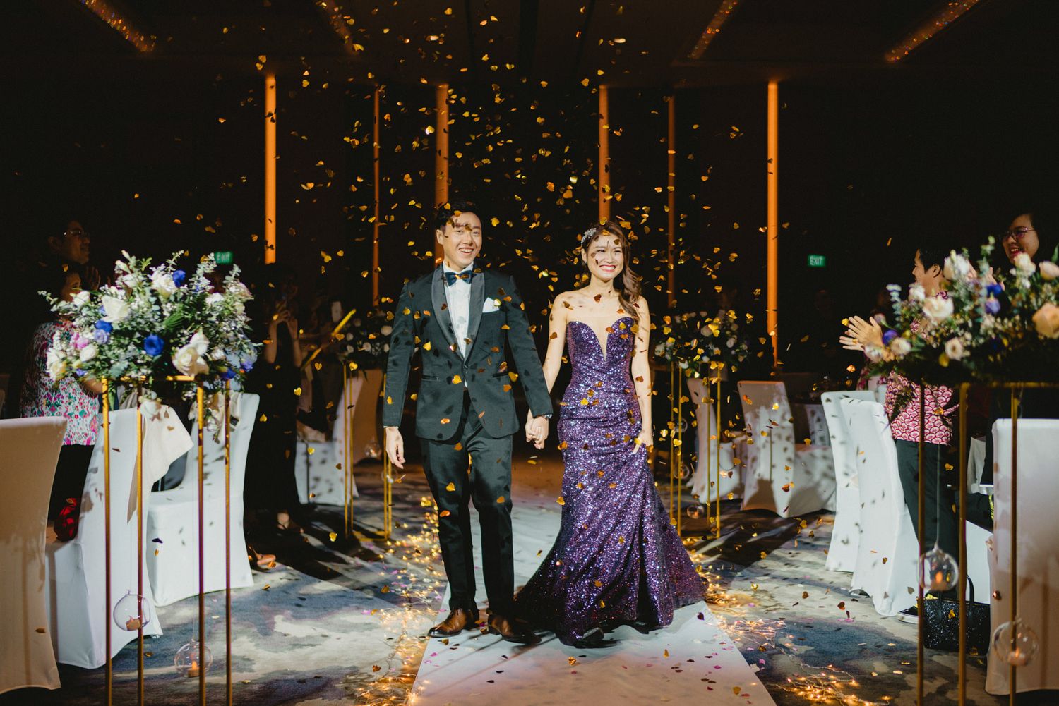 Couple in formal attire walks down wedding aisle amid golden confetti in elegant venue.