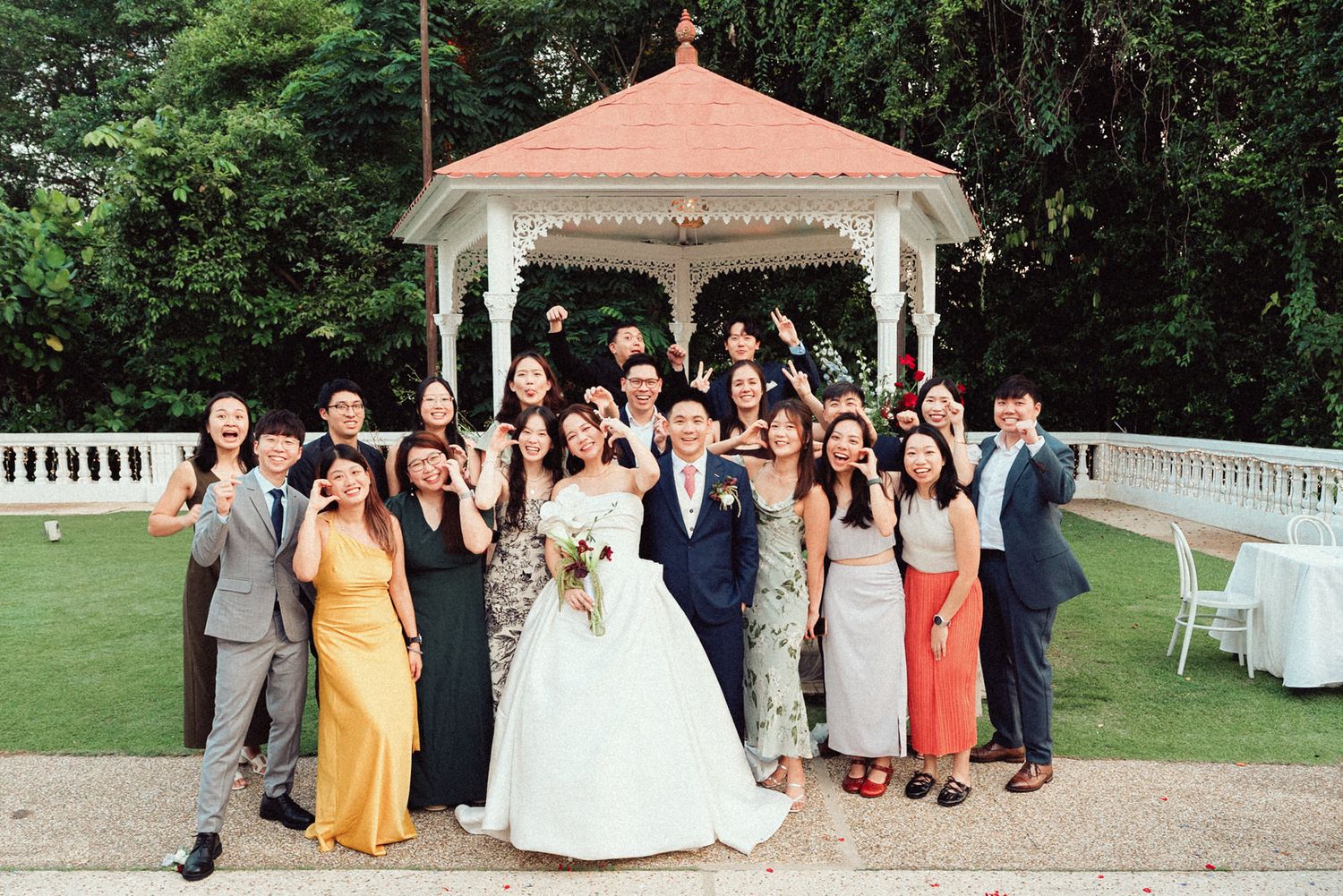 Wedding party poses outdoors near gazebo and garden during summer celebration.