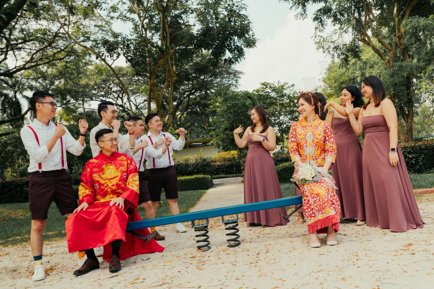 Chinese wedding party plays on a seesaw in a park, bride in red qipao with bridesmaids and groomsmen.