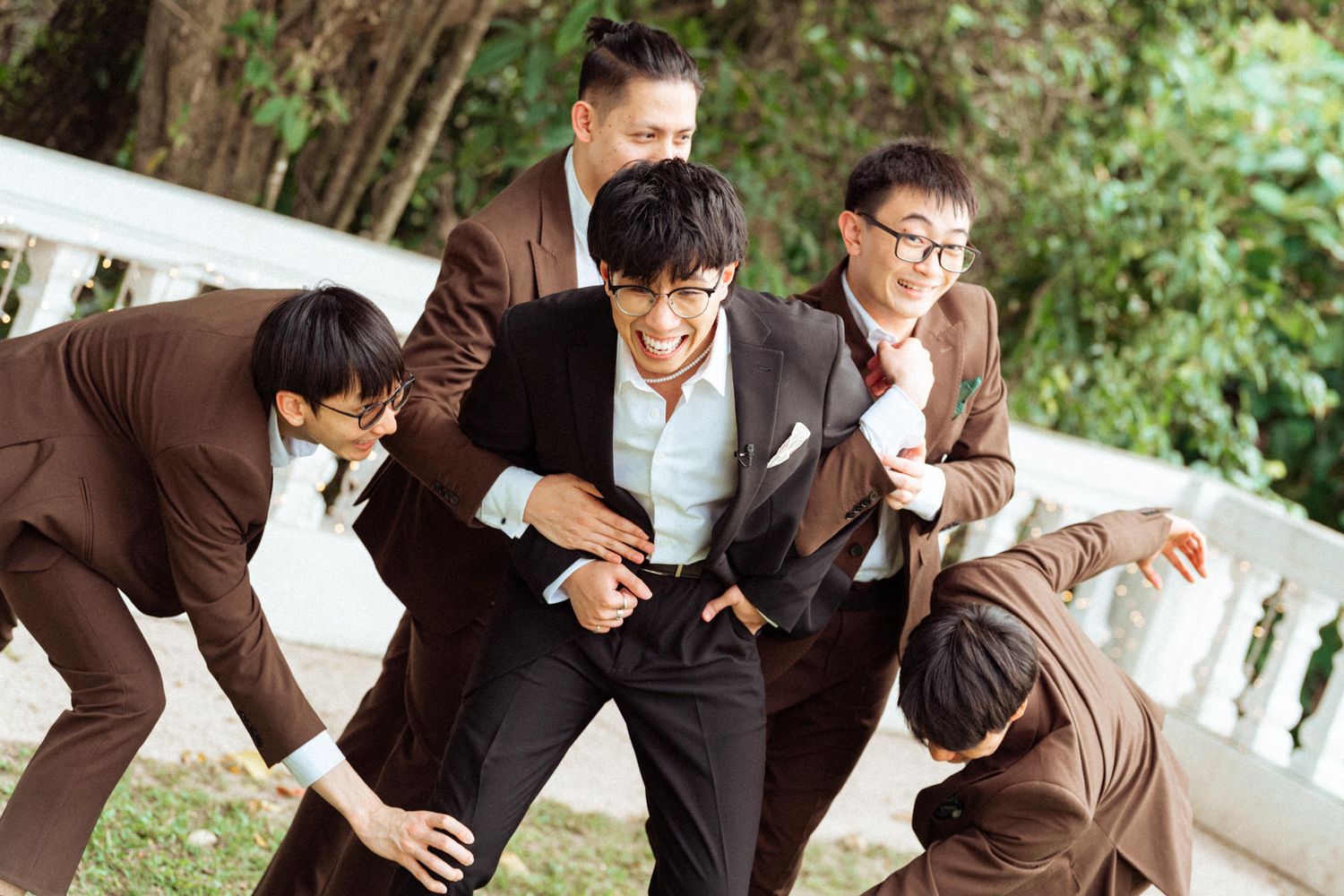 Group of five men in brown suits playfully posing outdoors, laughing and leaning together in a fun candid moment.