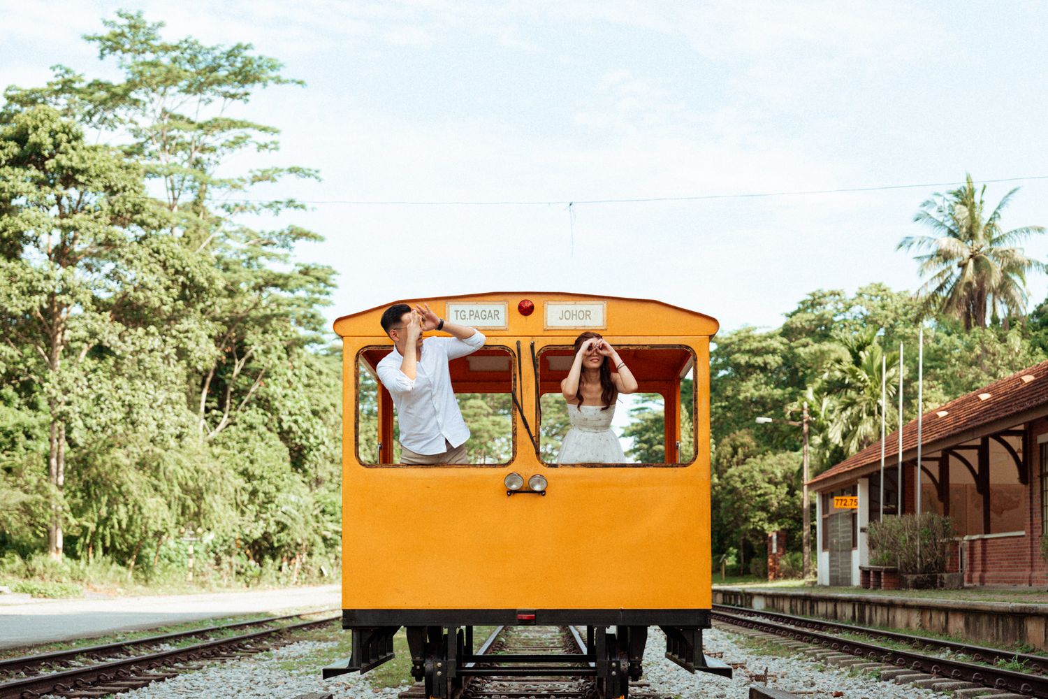 Two people waving from a bright orange train car on tracks surrounded by lush tropical trees.