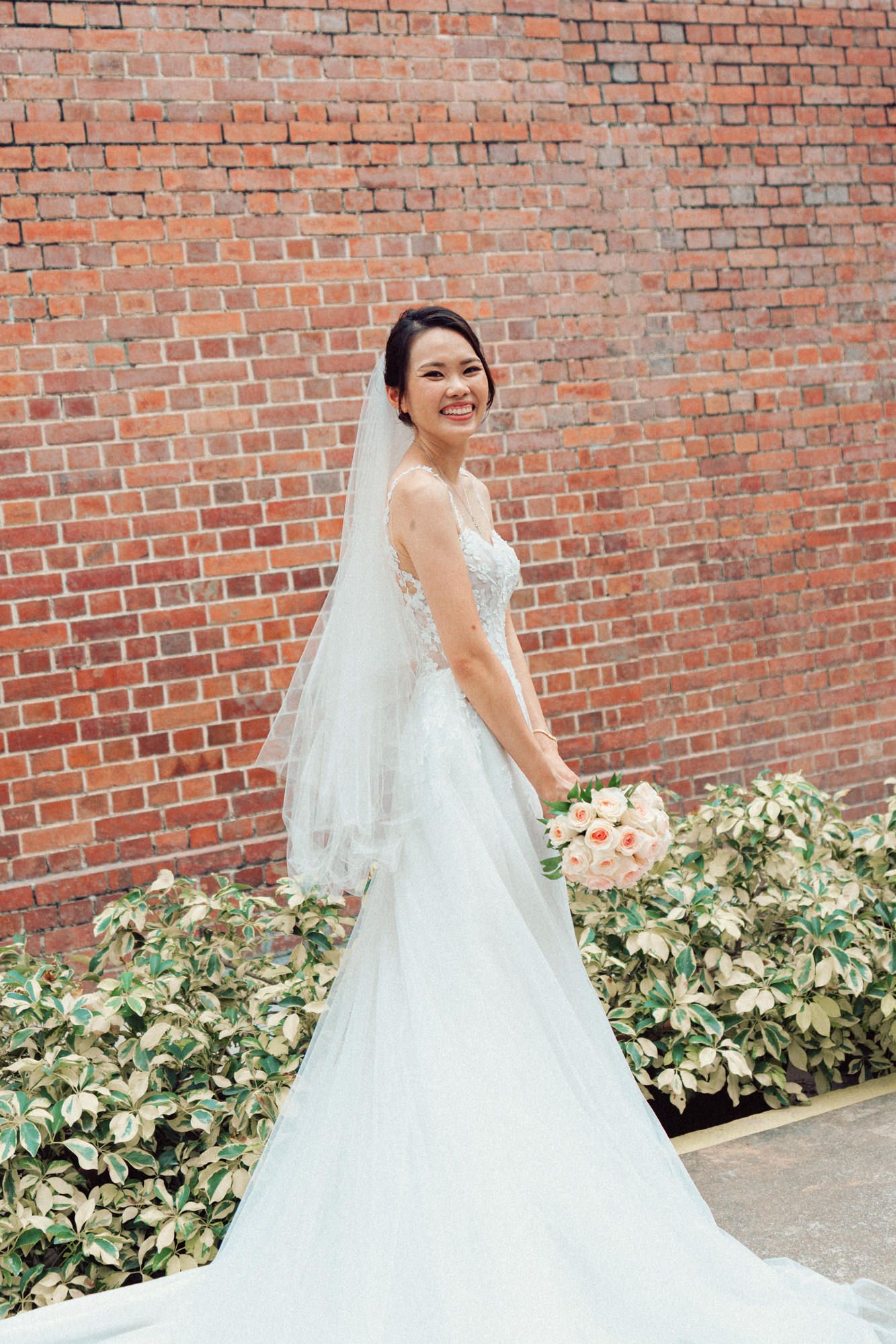 Elegant bride in lace wedding gown poses against brick wall with floral arrangements.