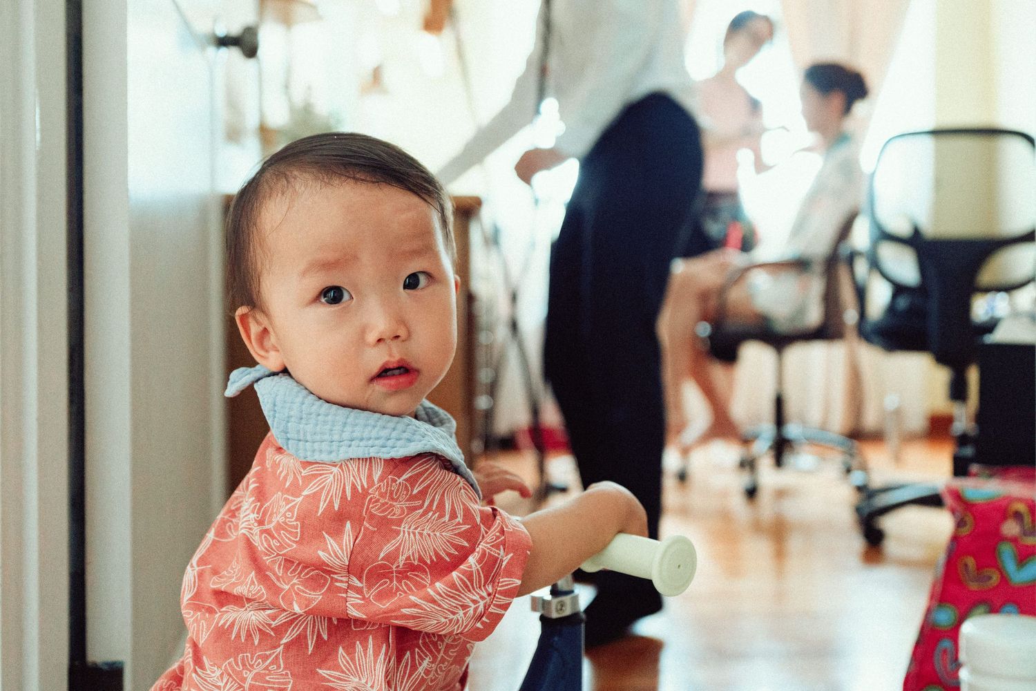 Young child in pink floral outfit sitting in foreground with people visible in background.