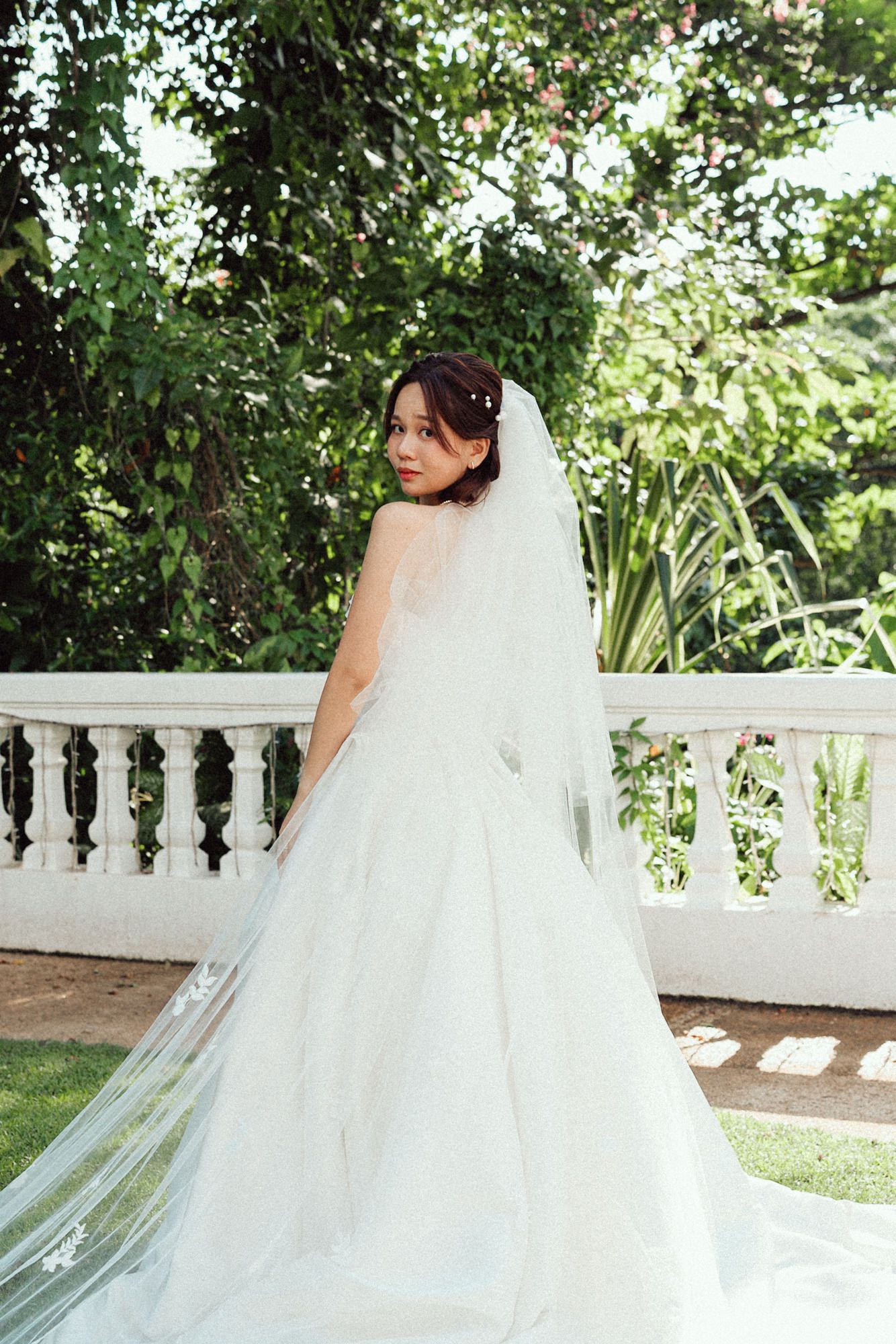 A flowing white wedding dress with a long veil captured in a garden setting against a white balustrade railing.
