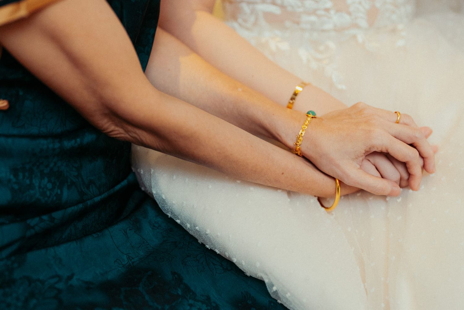 Close-up of two women holding hands, one wearing a gold bangle bracelet, in elegant teal and white dresses.