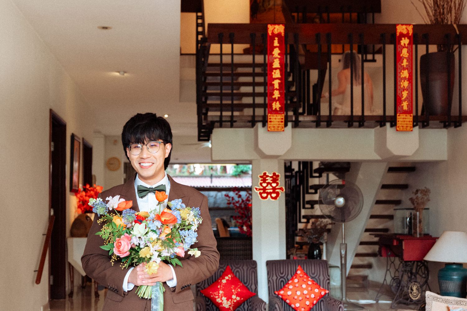 Young groom in brown suit holding colorful bouquet smiling at traditional Chinese wedding ceremony decoration.