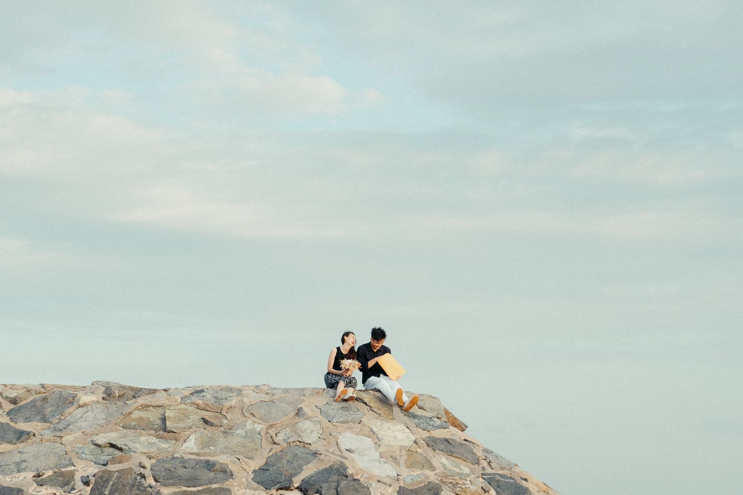 A couple sits together on coastal rocks overlooking the ocean.