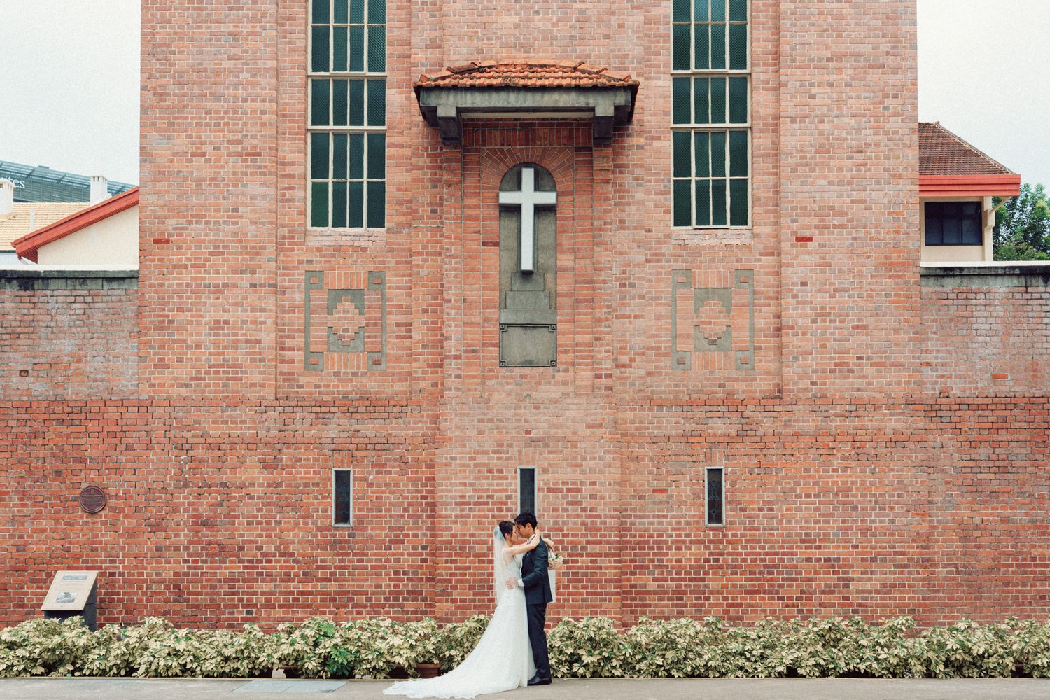 Wedding couple poses in front of brick church with cross and tall windows.
