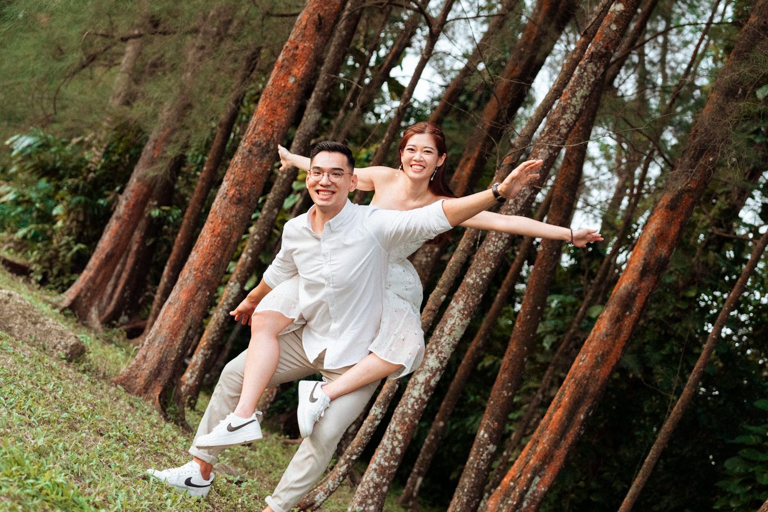 Playful couple photoshoot with arms spread wide in a pine forest.