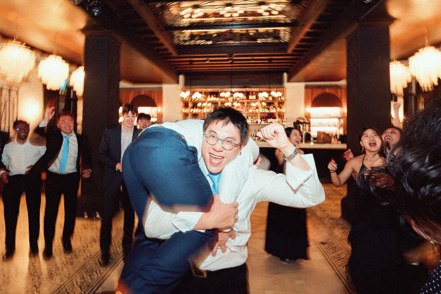 Wedding guests dance and celebrate together on an illuminated dance floor at an indoor reception venue.