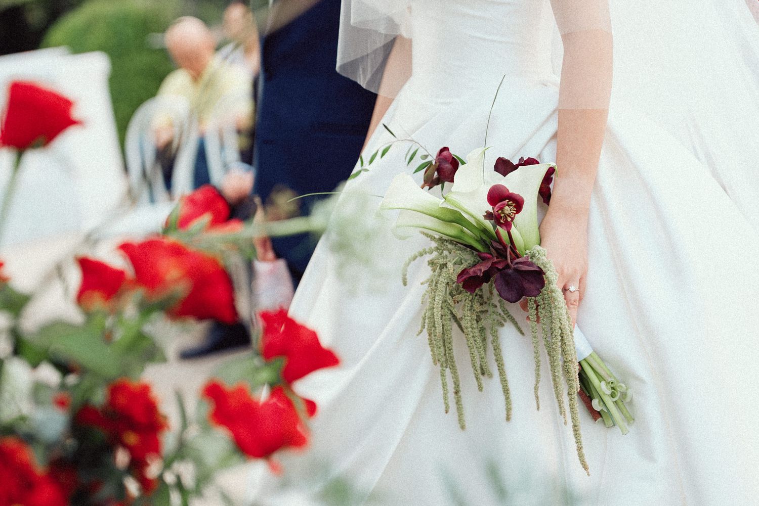 Bridesmaids in white dresses holding vibrant red rose bouquets walk together in an outdoor wedding ceremony.