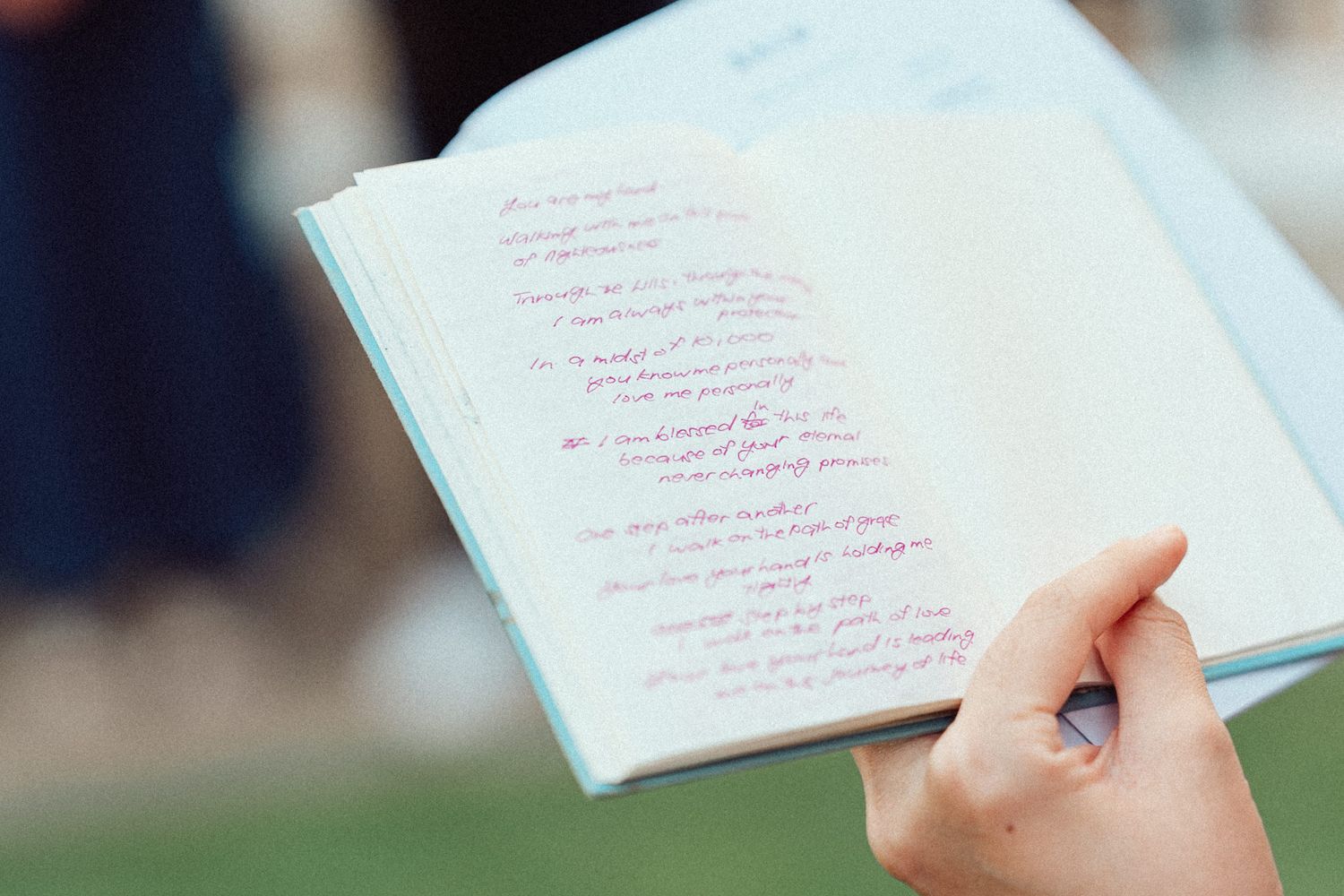 Hand holds open book pages showing handwritten text in red ink against blurred background.