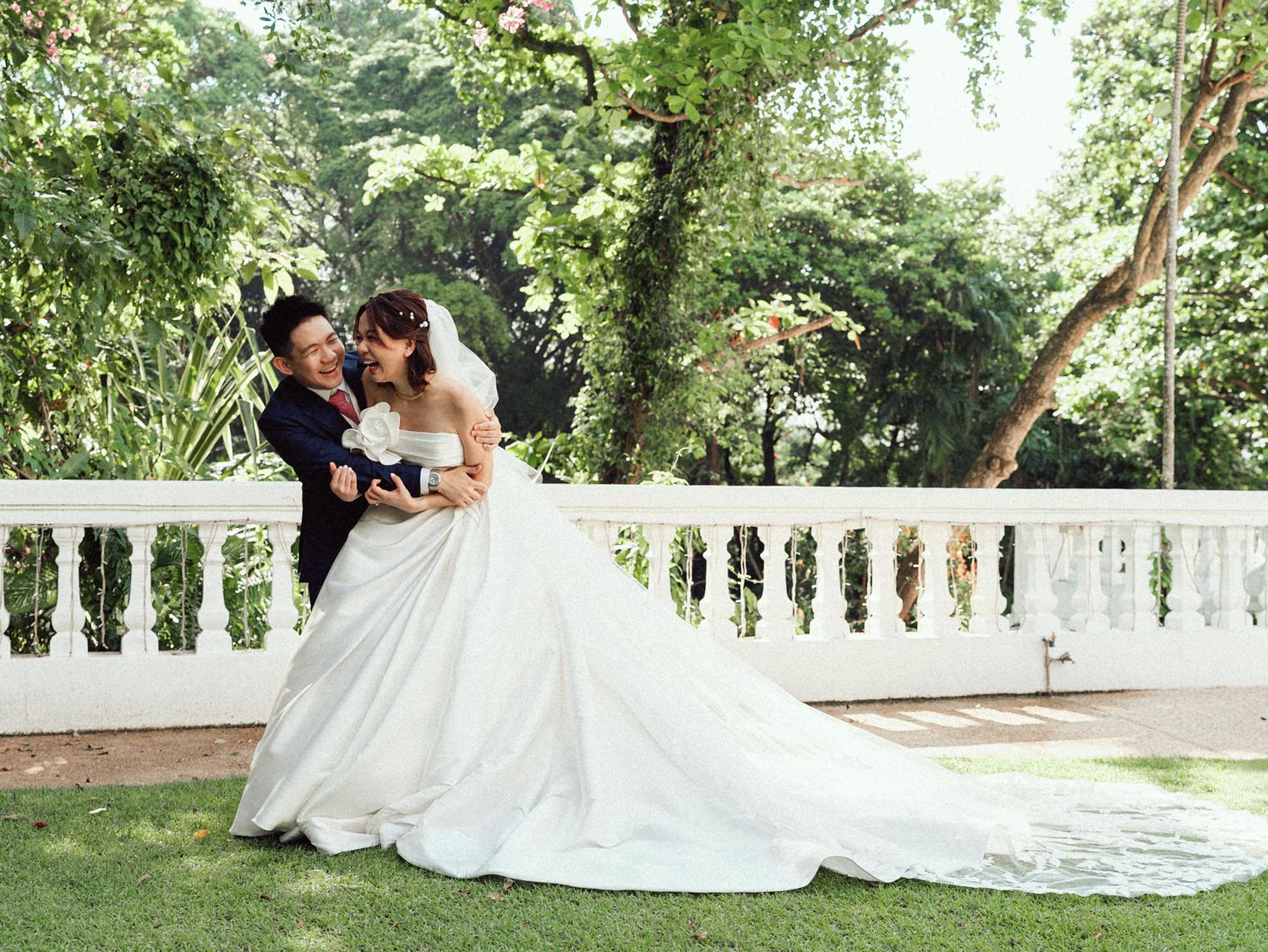 A couple in wedding attire shares an intimate moment together on a white bench in a lush garden setting.