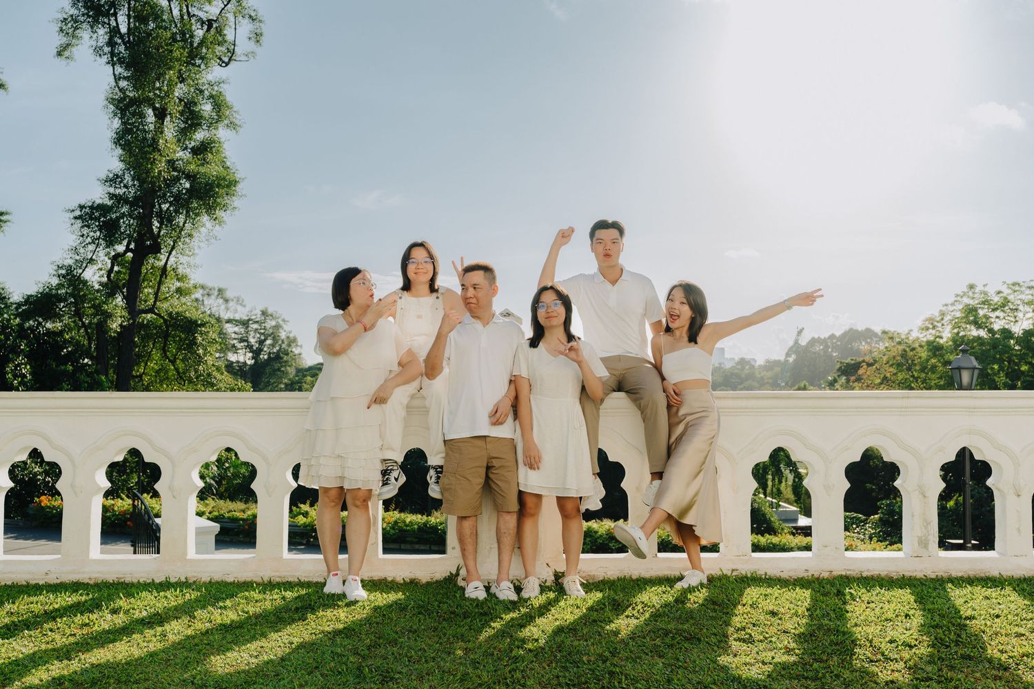 A joyful group in light-colored outfits poses together on a sunny terrace with greenery and architectural details.