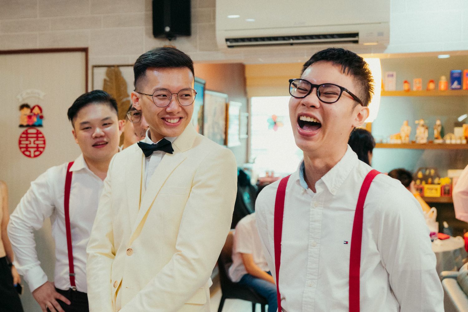 Three young men laughing and celebrating at a wedding, groom in white tuxedo with bow tie, groomsmen in red suspenders.