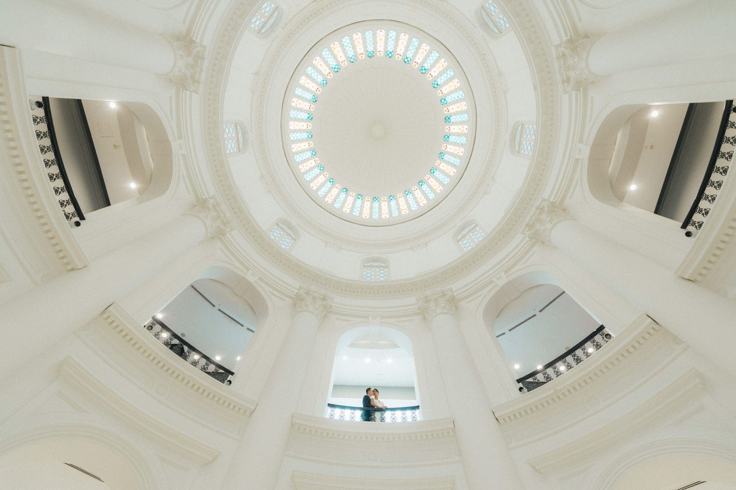 Elegant white domed rotunda interior with ornate architecture and a couple standing beneath a colorful stained glass ceiling.