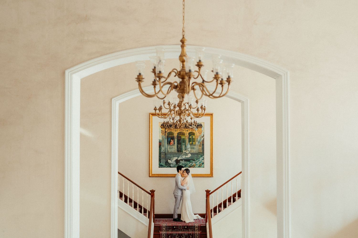 Bride and groom embracing on elegant staircase under gold chandelier in a grand venue.