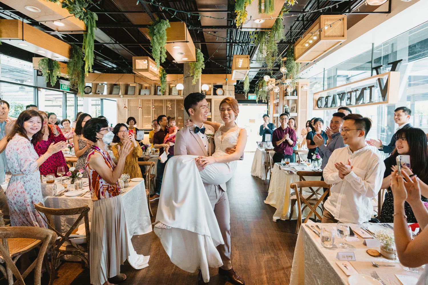 Joyful wedding reception in a modern restaurant with guests applauding the bride and groom surrounded by greenery.